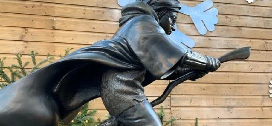 Bronze statue of a person riding a broomstick in front of a wooden wall with decorative elements on Harry Potter Filming Locations Tour