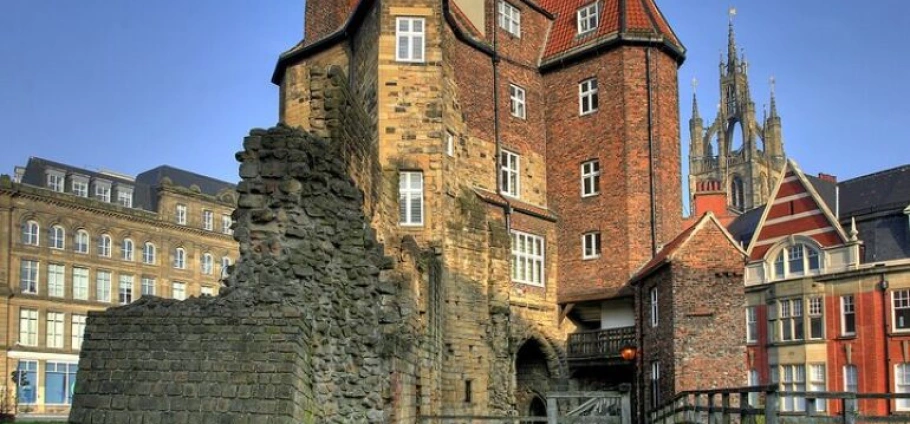 Newcastle Castle | Old brick and stone castle with various buildings in the background, including a church with a tall spire.