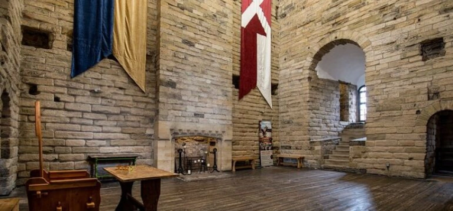 Newcastle Castle | A large stone-walled room with tall ceilings, showcasing a medieval interior with tapestries hanging on the walls, a central fireplace, wooden benches, and arched doorways and windows.