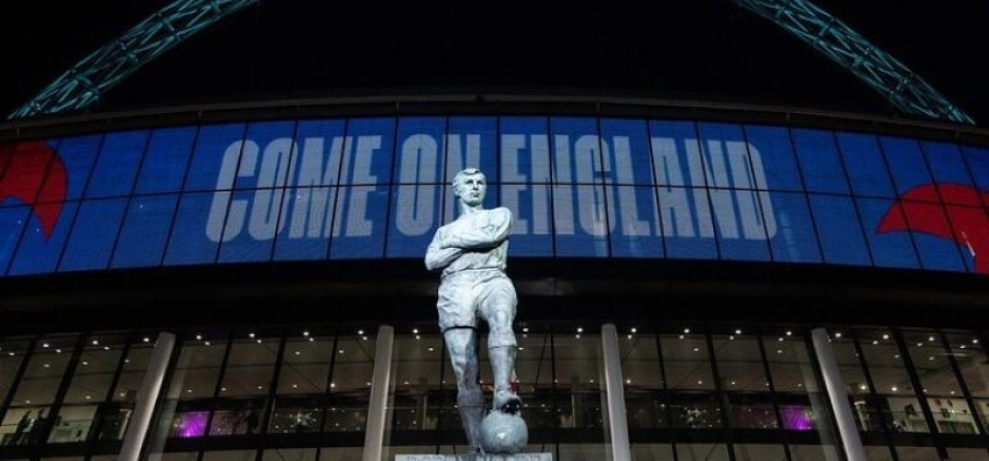 Statue of a football player in front of Wembley Stadium with "Come On England" displayed on the screen.