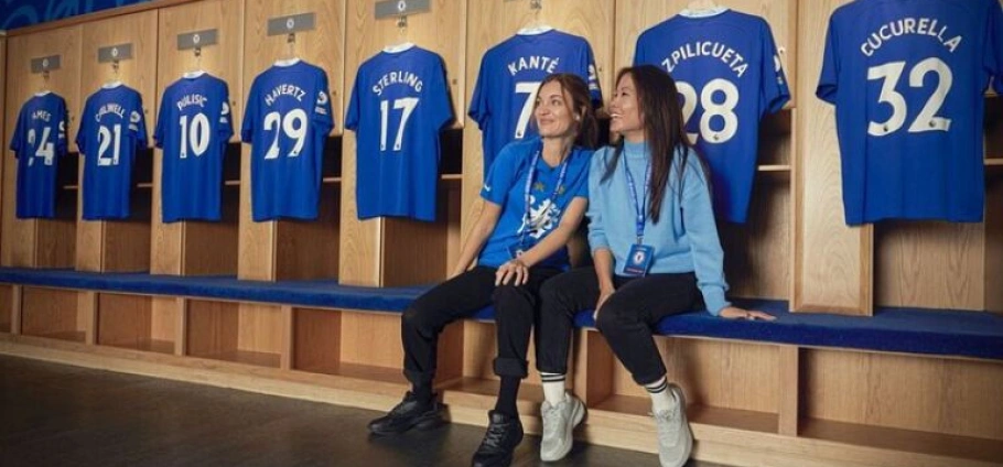 Two women sitting in a football locker room with blue jerseys hanging behind them on the London Chelsea FC Stadium Tour.