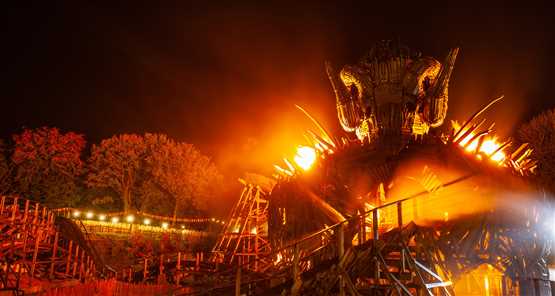 Nighttime view of an illuminated wooden roller coaster featuring a large, fiery effigy with a horned head.