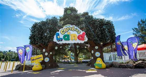 Entrance to CBeebies Land with colourful signage and trees, flanked by CBeebies characters and welcome flags.