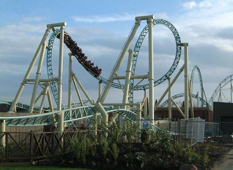 A roller coaster with teal tracks and beige supports featuring a large loop and a train of riders descending a steep drop against a partly cloudy sky