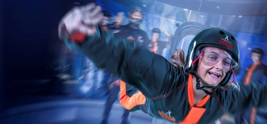 Person indoor skydiving in a wind tunnel wearing safety gear and helmet, with instructors in the background