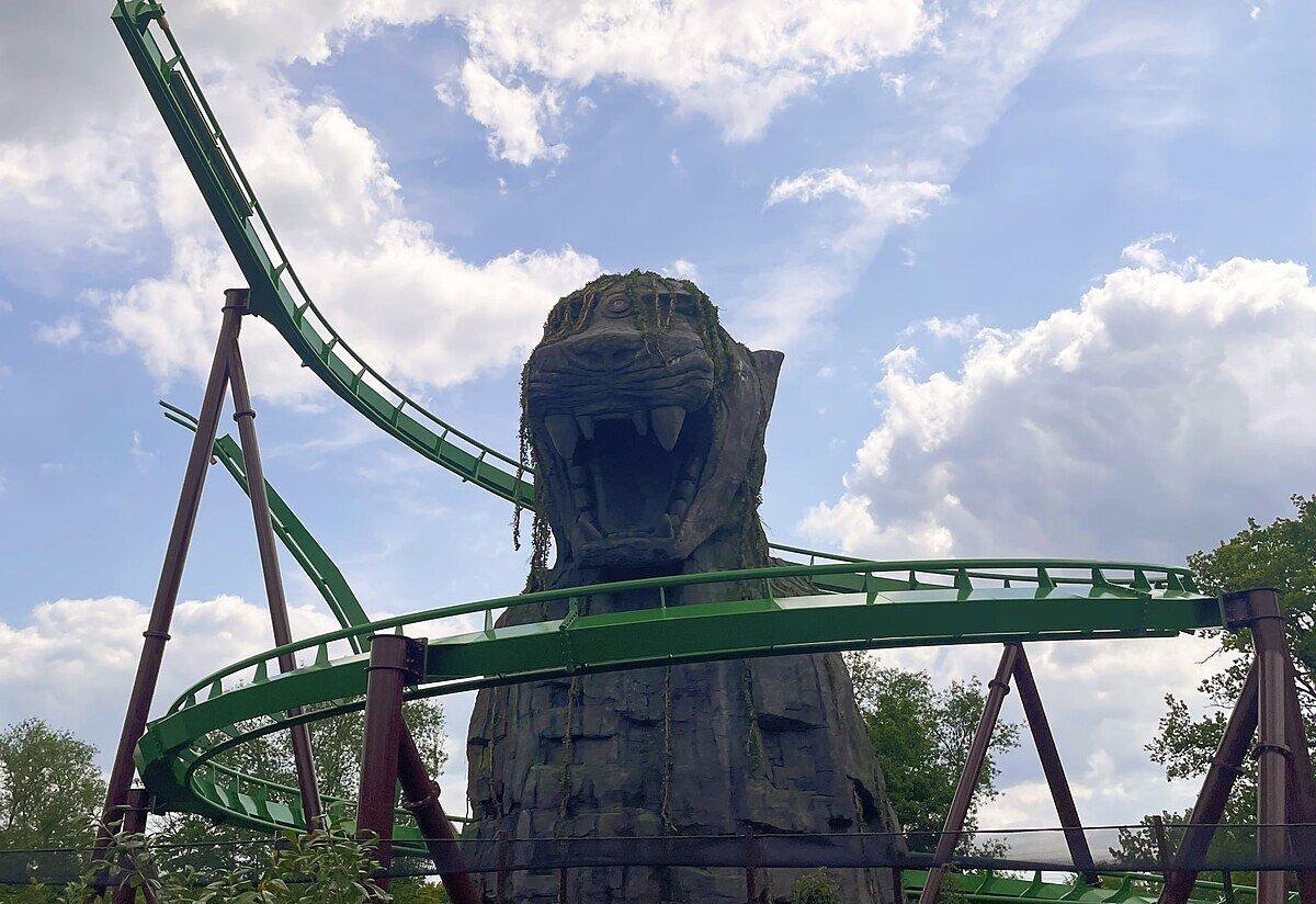 Green roller coaster track curving around and over a large statue of a roaring tiger head with moss hanging from it against a partly cloudy sky.