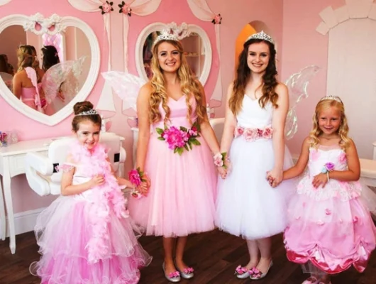 Four girls dressed as fairies and princesses in a pink-themed room with mirrors and vanity tables.