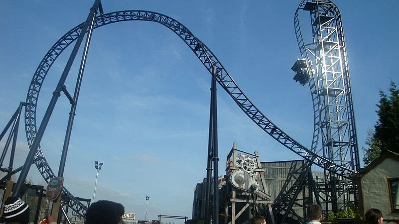 Large roller coaster structure with a steep vertical drop and looping track against a clear blue sky, with some people visible below