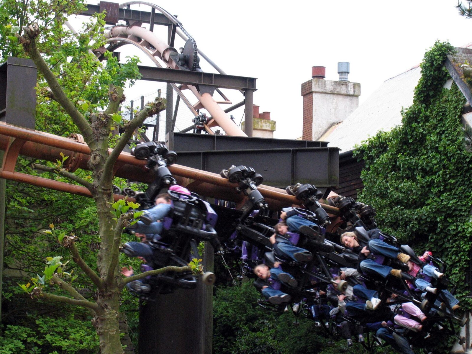 Roller coaster with riders seated and secured on a suspended track, turning sharply near a tree, surrounded by greenery and buildings with chimneys in the background.
