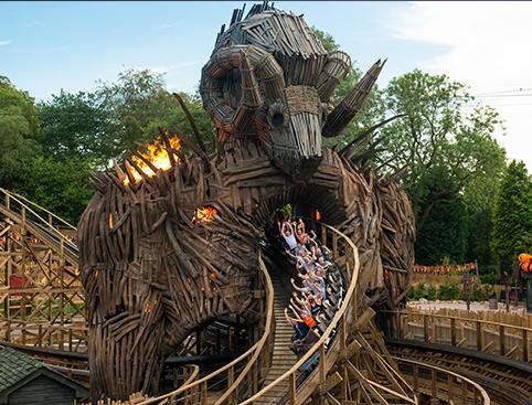 A wooden roller coaster with a large wooden figure and flames, surrounded by greenery.