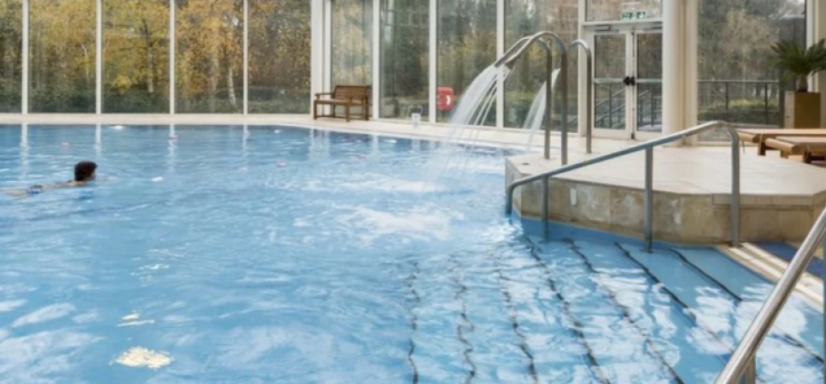 Indoor swimming pool with a person swimming, featuring large windows, water fountains, and poolside seating at Birmingham PURE Spa.