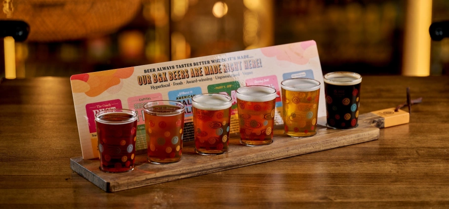 A flight of craft beers on a wooden tray with a colorful menu in the background, displayed on a bar counter. Craft Beer Experience