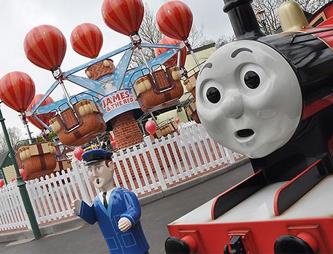 A theme park ride featuring hot air balloon baskets and a "James the Red Engine" sign, with a statue of a train and a conductor figure in the foreground.