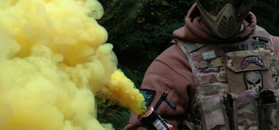 Paintball player in camouflage gear holding a yellow smoke grenade in a forest setting at Group Paintball Experience Birmingham