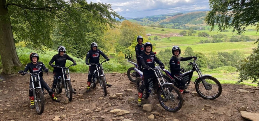 Group of six people in motorcycle gear with trials bikes on a scenic hillside with green fields and rolling hills in the background.