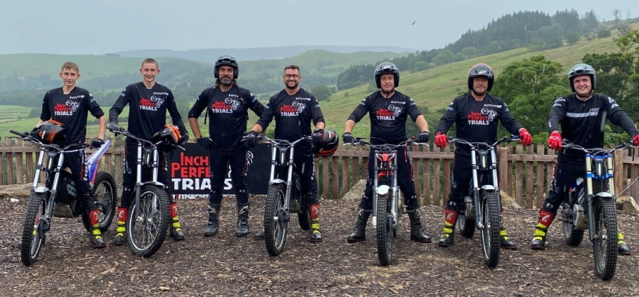 Group of seven men standing with trial bikes, wearing matching "Inch Perfect Trials" jerseys, outdoors with rolling hills in the background.