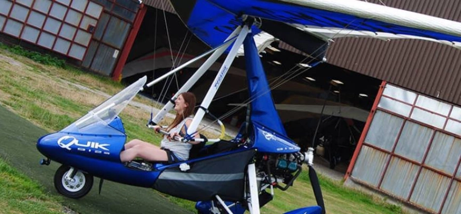 A woman sitting in the cockpit of a blue microlight aircraft parked near a hangar.