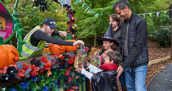 LEGOLAND Windsor Resort | Family enjoying Halloween event with children in costumes receiving candy from a security guard at a decorated booth in a park.