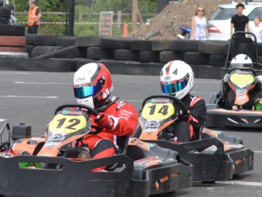 Go-kart racers competing on an outdoor track with spectators in the background at Lichfield Go Kart Driving