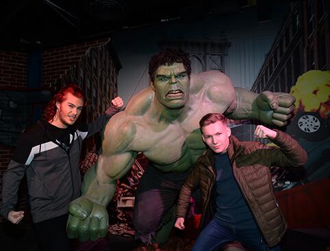 Two people posing with a giant green Hulk statue in a superhero-themed setting.