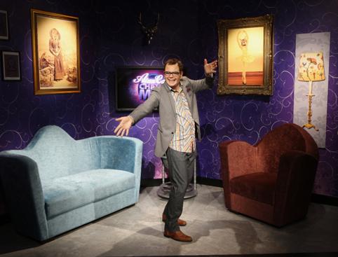 A person standing in a purple-themed studio with two chairs, dressed in a suit with an open arms gesture, and smiling.