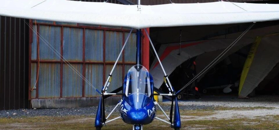 Small blue ultralight aircraft in front of a hangar.