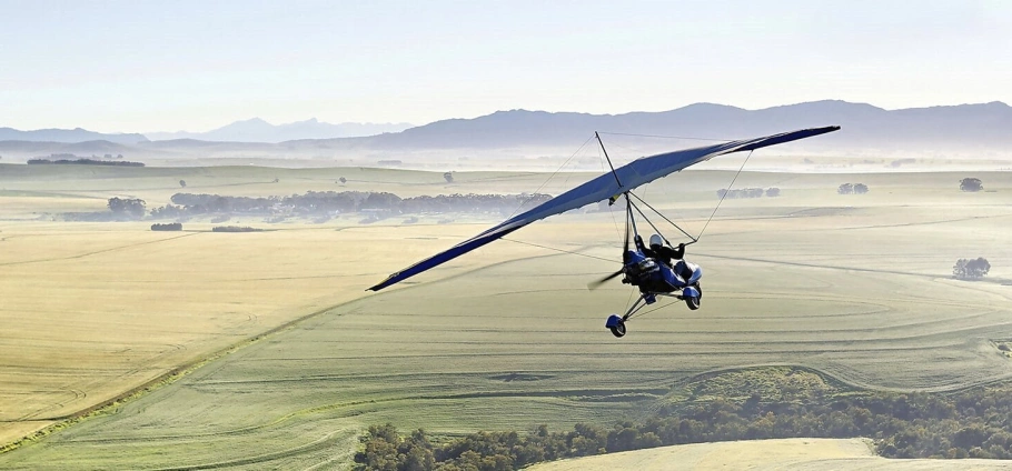 Microlight aircraft flying over a vast agricultural landscape with fields and distant mountains in the background.