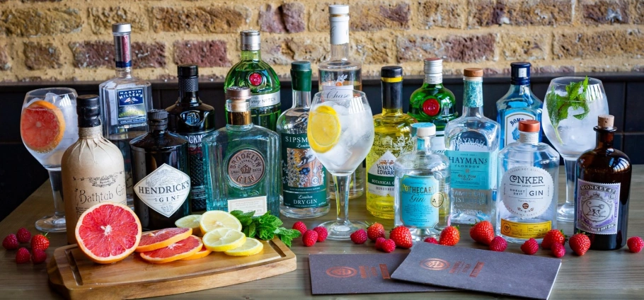 Assorted gin bottles and cocktails with garnishes on a table in front of a brick wall.