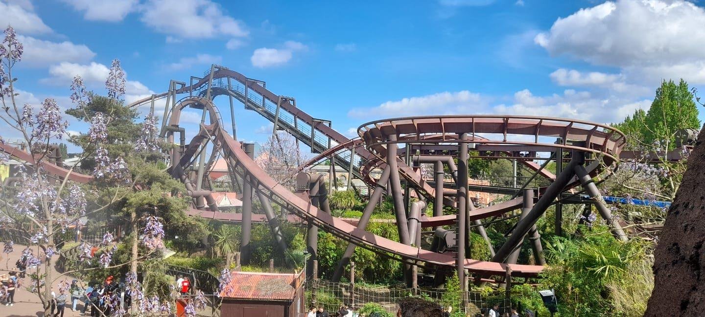 A roller coaster track with rust-coloured rails twisting and looping above green foliage, with people walking below and a partly cloudy blue sky overhead.