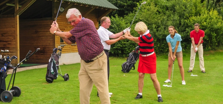 PGA Professional 30 Minute Golf Lesson | Group of people taking golf lessons on a green field with golf clubs and bags nearby.