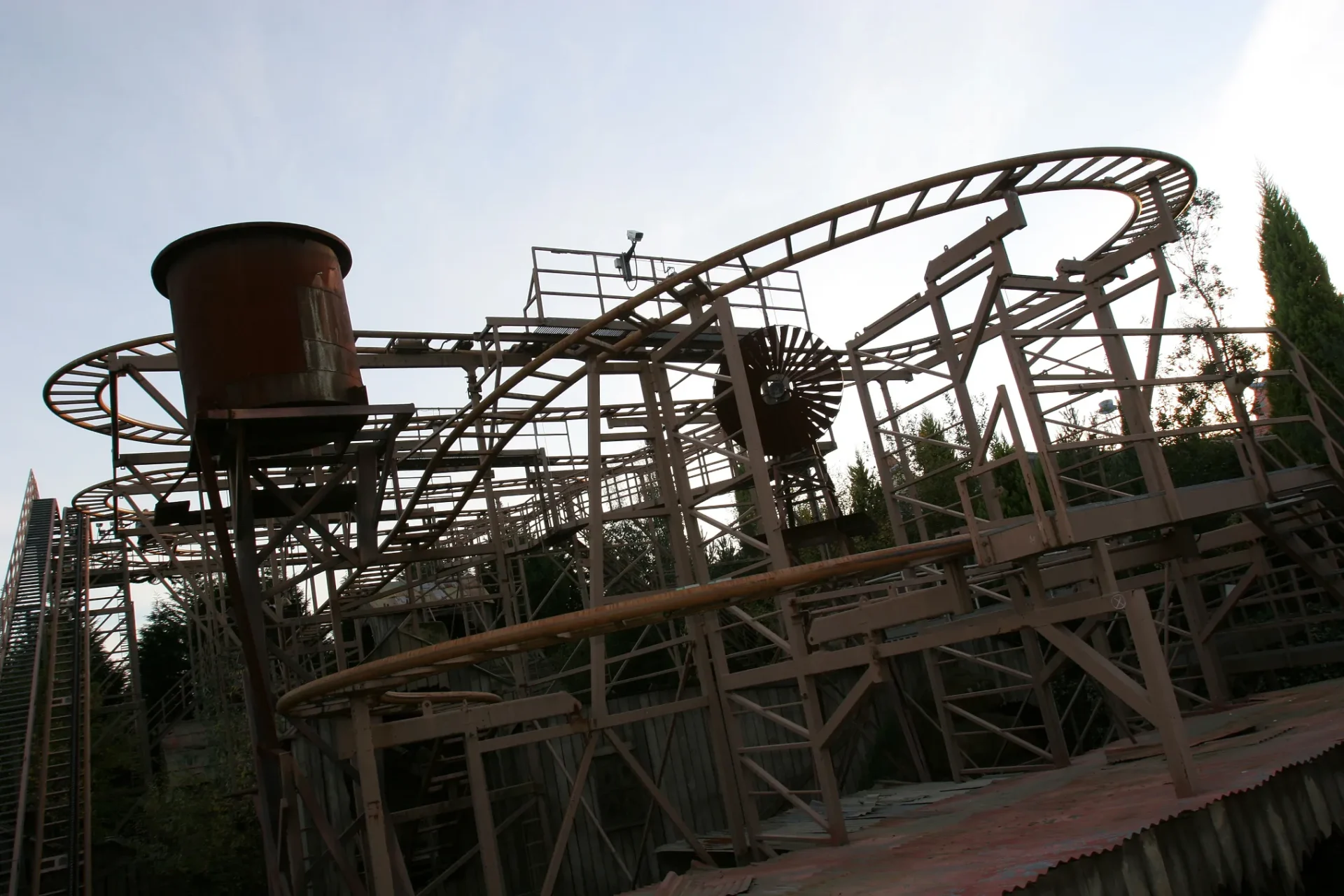 Rusty, abandoned roller coaster structure with a dark water tower and curved tracks against a cloudy sky