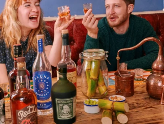 Two people enjoying drinks at a bar with various bottles of alcohol and a small distillation setup on the table.