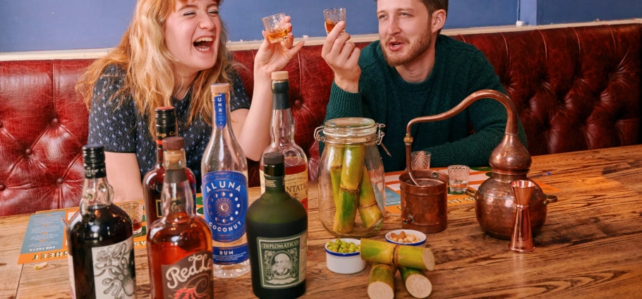 Two people enjoying drinks at a bar with various bottles of alcohol and a small distillation setup on the table.