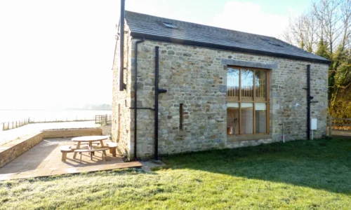 Stone cottage with large wooden windows, outdoor picnic bench, and surrounding fields and trees.