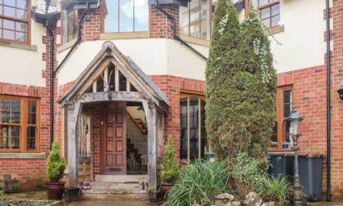 Front entrance of a two-storey brick house with timber-framed porch, central wooden door, windows with wooden frames, green plants, and a streetlamp.