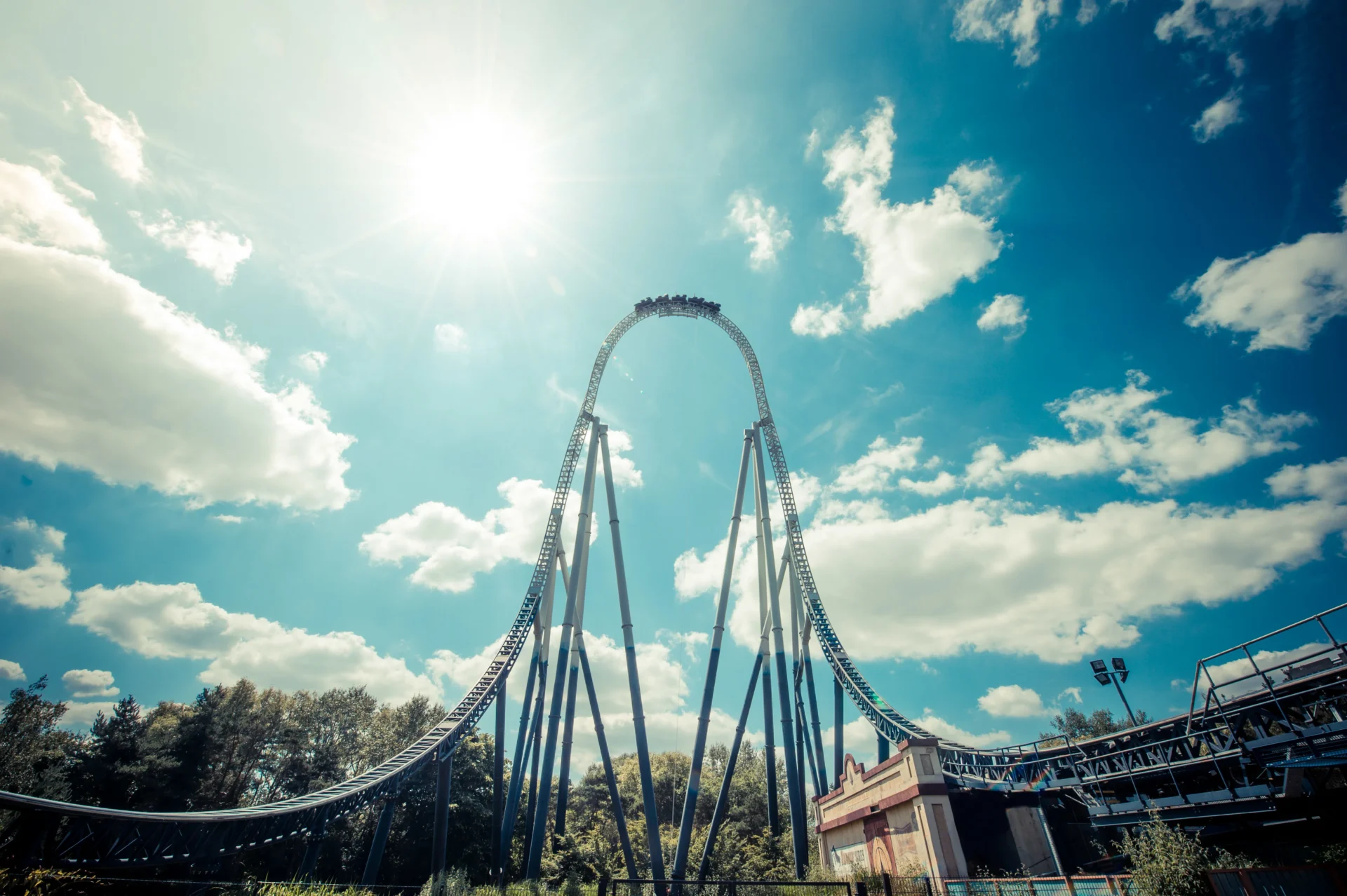 Roller coaster with a steep loop towering against a bright blue sky with scattered clouds and sun shining brightly above, surrounded by trees and a building near the coaster's base.