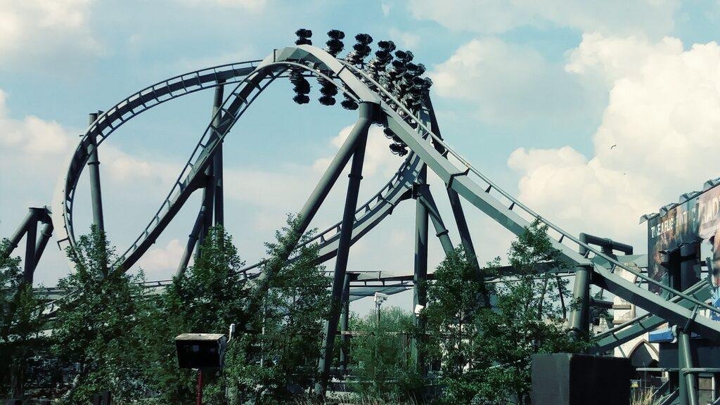 Inverted roller coaster train with riders going through a loop on a metal track set against a partly cloudy sky with green trees in the foreground.