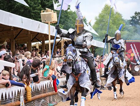 Warwick Castle Tickets | Two knights on horseback, wearing armour and holding lances, participate in a jousting tournament with a crowd of spectators in the background.