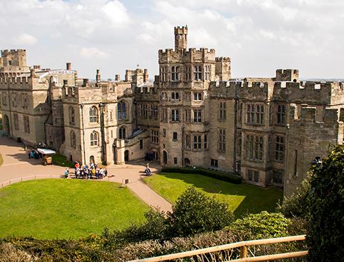 Warwick Castle Tickets | Medieval castle with multiple towers, a large central courtyard, and groups of people walking and sitting on the grass.