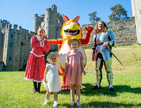 Warwick Castle Tickets | Two children pose with a person in dragon costume, a knight, and a woman in historical attire in front of a castle.