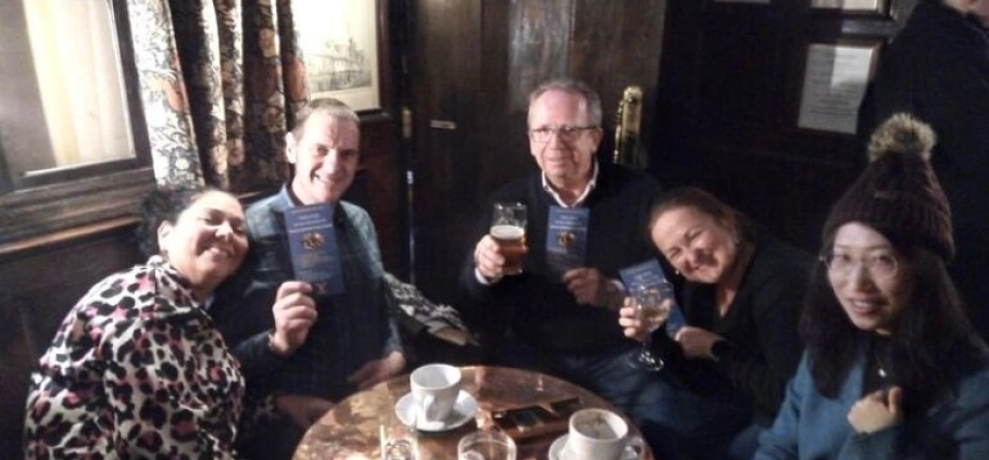 Five people sitting at a pub table holding drinks and booklets, smiling at the camera.