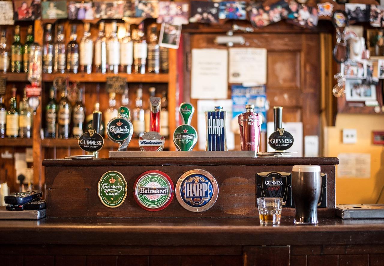 Bar counter with beer taps for Carlsberg, Guinness, Heineken, Budweiser, Harp, and Tetley's. Background includes shelves stocked with bottles and photos.