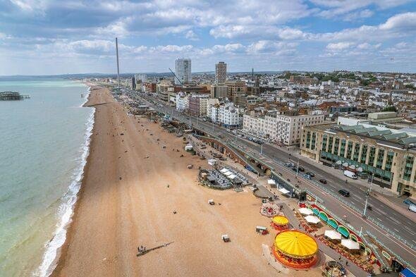 Brighton Beach: A wide aerial view of a sandy beach with people scattered along the shore, a coastal road running parallel to the beach, rows of buildings and high-rises in the background, and a yellow carousel with colourful stalls near the bottom right corner.