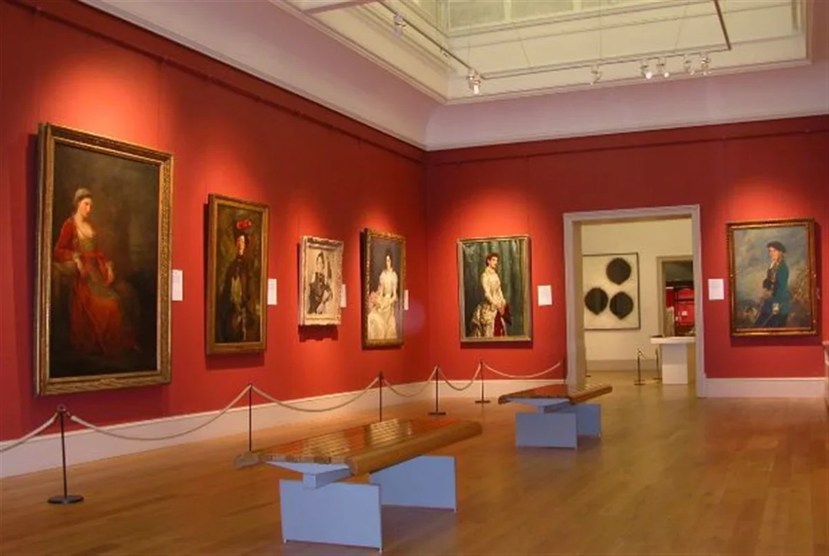 Interior of an art gallery with red walls displaying seven framed classical portraits and wooden benches on a polished wooden floor under a high ceiling with skylights.