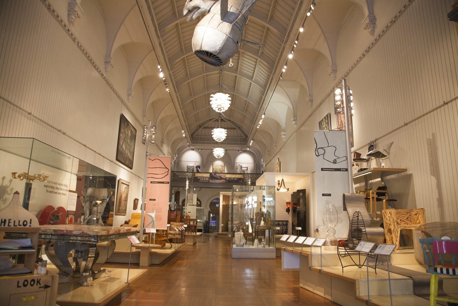 Interior view of an art gallery with a high, vaulted ceiling and wooden floor, displaying various modern art pieces, sculptures, and furniture along the walls and enclosed in glass cases in the centre, with warm overhead lighting.