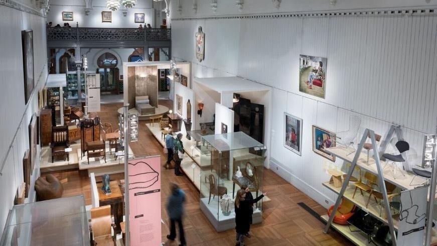 Interior view of a museum gallery featuring various furniture pieces, display cases, and framed photographs on the walls, with several visitors observing the exhibits.