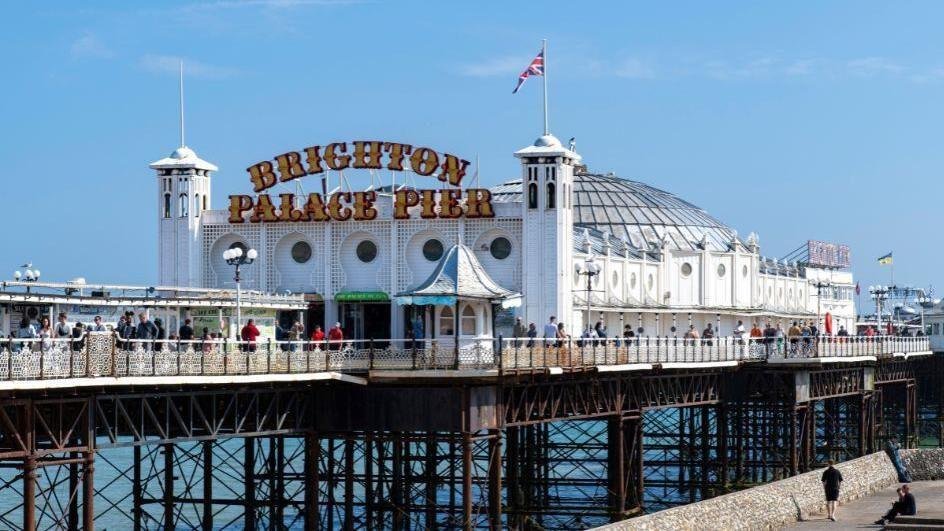 Brighton Palace Pier in the United Kingdom with its white building and large sign, people walking along the pier, and the sea underneath on a clear sunny day