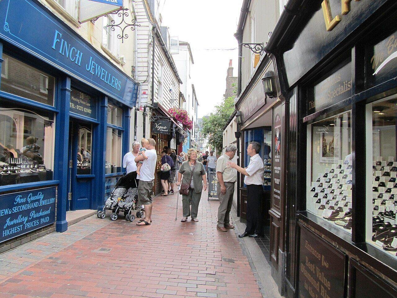 The Lanes, Brighton: Narrow pedestrian street with brick pavement, lined with jewellery shops on both sides, people walking and looking into shop windows on a cloudy day