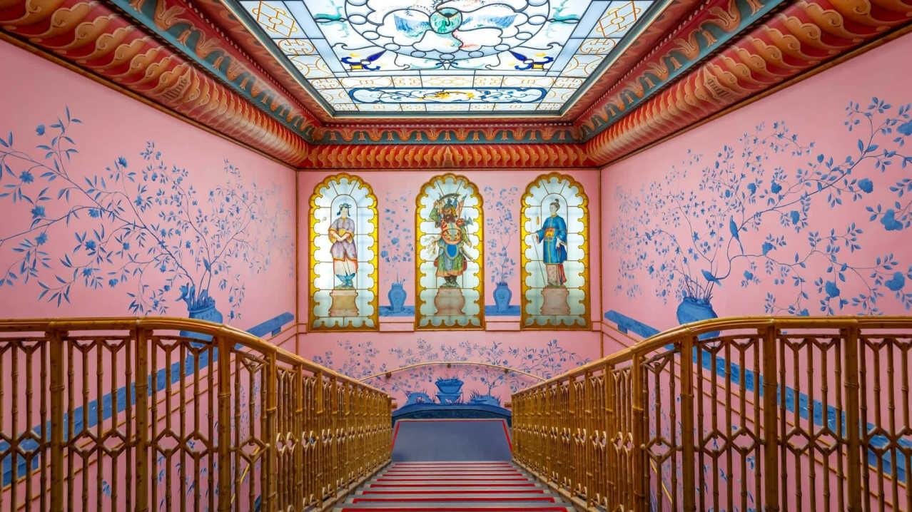 Discover the Royal Pavilion: Staircase with ornate wooden railings leading down to a landing with pink walls decorated with blue floral and bird motifs, three arched stained glass windows featuring figures, and a ceiling with a stained glass panel depicting a floral design.