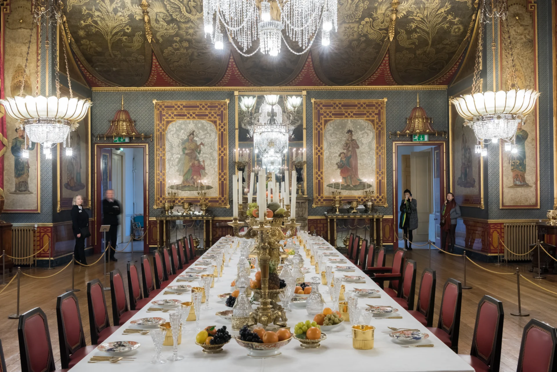 Grandiose dining room with an ornate long table set with fine china, crystal glasses, gold cutlery, and fruit bowls, flanked by red-upholstered chairs, under crystal chandeliers and lavishly decorated walls with large framed paintings and intricate ceiling patterns, with a few people standing near doorways on each side of the room.
